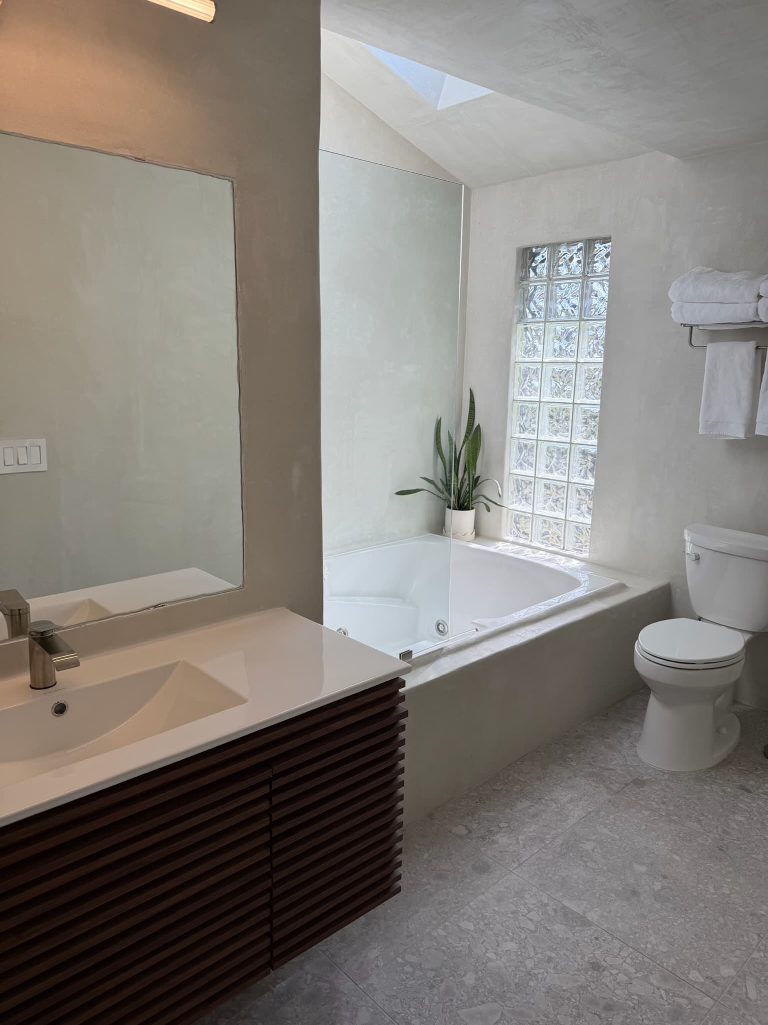 A soaking tub set beneath a skylight and glass-block window in a microcement bathroom with a walnut vanity.