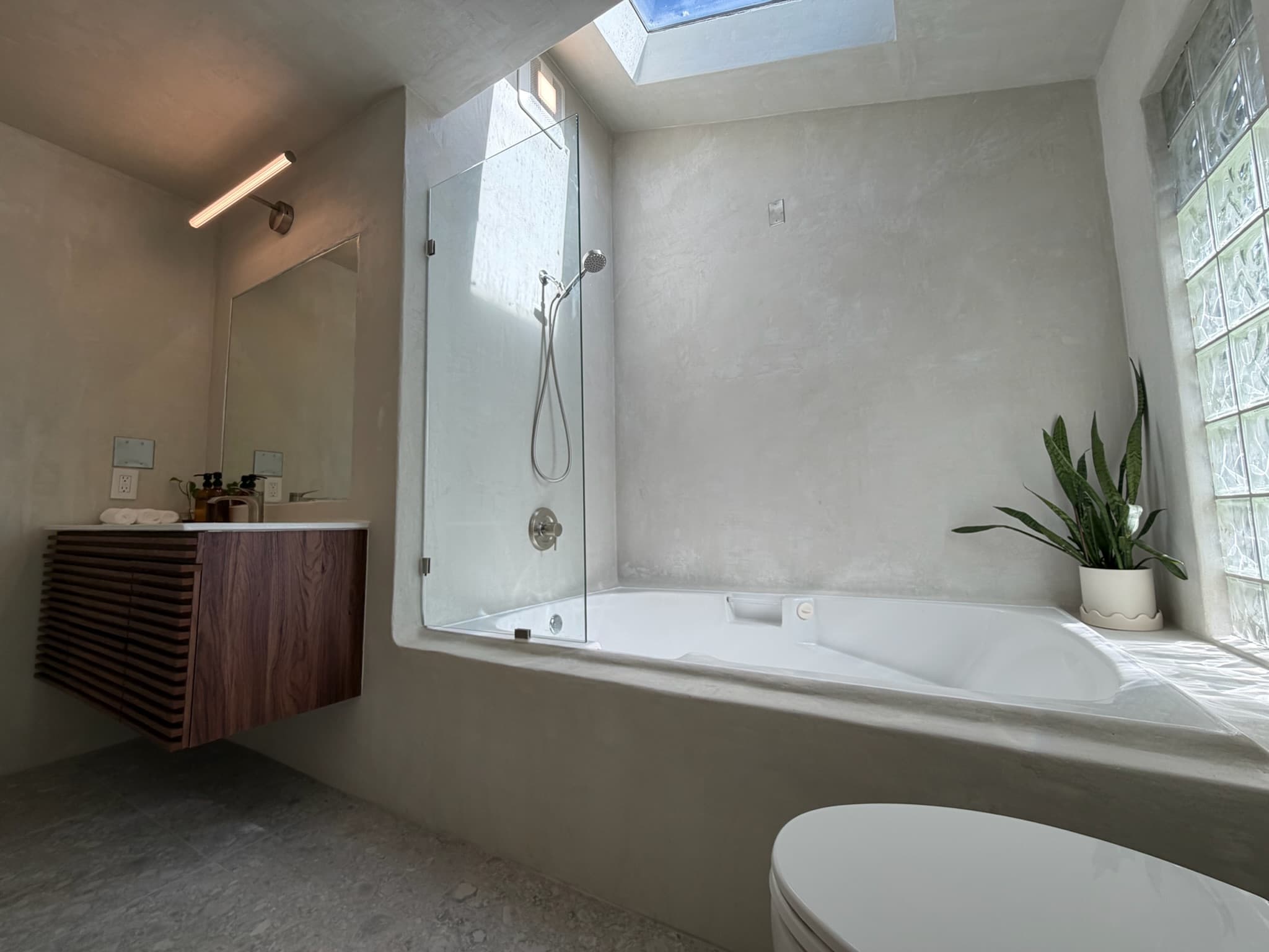 Wide view of a microcement spa bathroom with a glass shower panel, soaking tub, floating walnut vanity, and skylight.