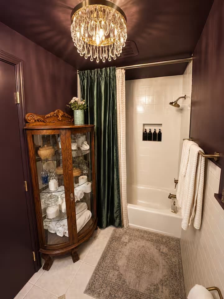 The same plum bathroom with a crystal chandelier, antique curio cabinet, and a green velvet shower curtain.