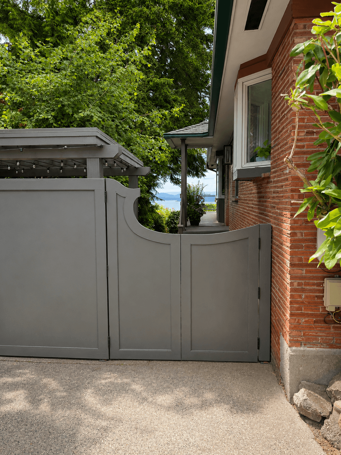 Close view of a gray custom gate with a scalloped top rail and a lit trellis cover next to a brick house.