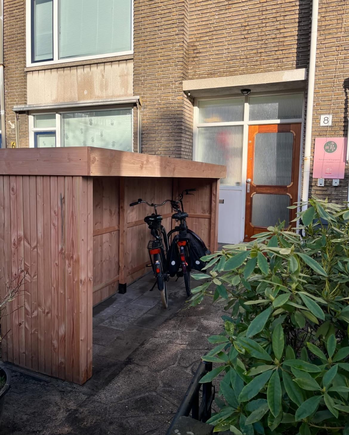 A cedar storage cover with a flat roof sheltering bikes against a brick house entry.